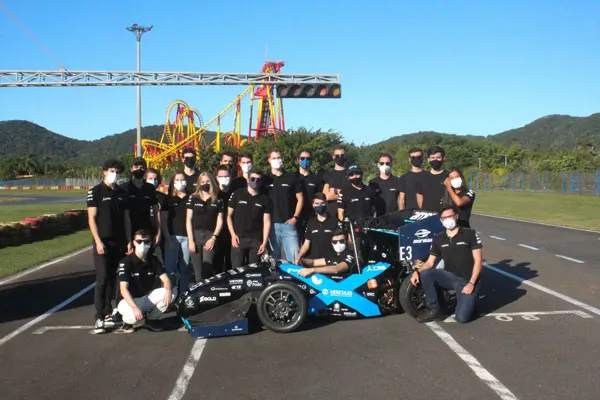 A group of young adults standing around an autonomous race car, on a race track, with a rollercoaster in the background.